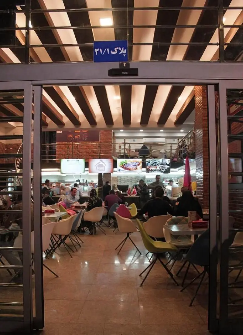 تصویری از ورودی و فضای داخلی شلوغ فست فود خانه برگر با مشتریانی که از غذا لذت می‌برند. محیطی دلنشین برای صرف ناهار و شام.