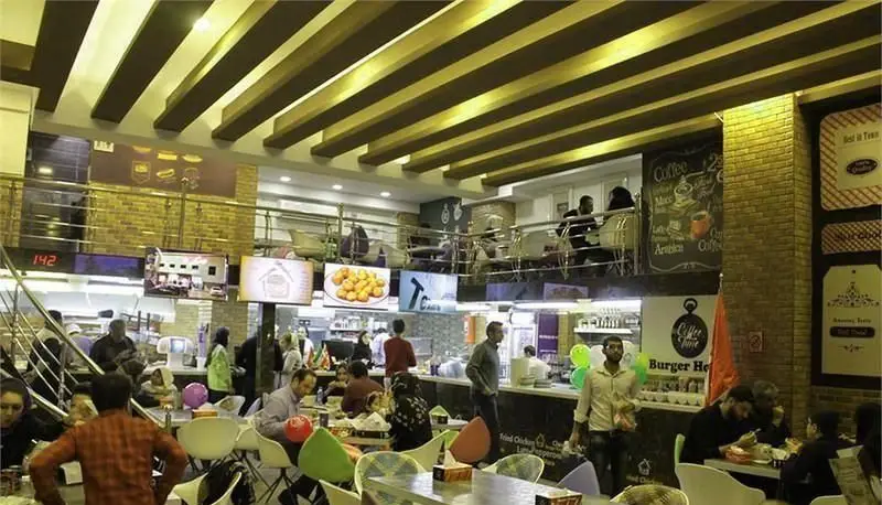 تصویری از داخل شلوغ و چندطبقه فست فود خانه برگر، نمایش مشتریان، میزها، پیشخوان و تابلوهای منوی دیجیتال.