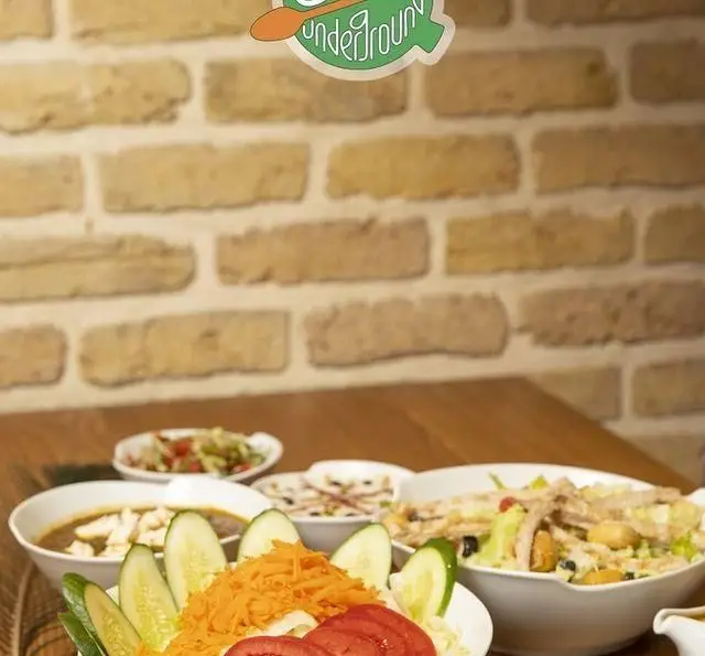 میز چوبی کافه رستوران زیرزمین با غذاهای متنوع شامل سالادهای رنگارنگ، سوپ و پیش‌غذا در پس‌زمینه آجری و لوگوی زیرزمین.
