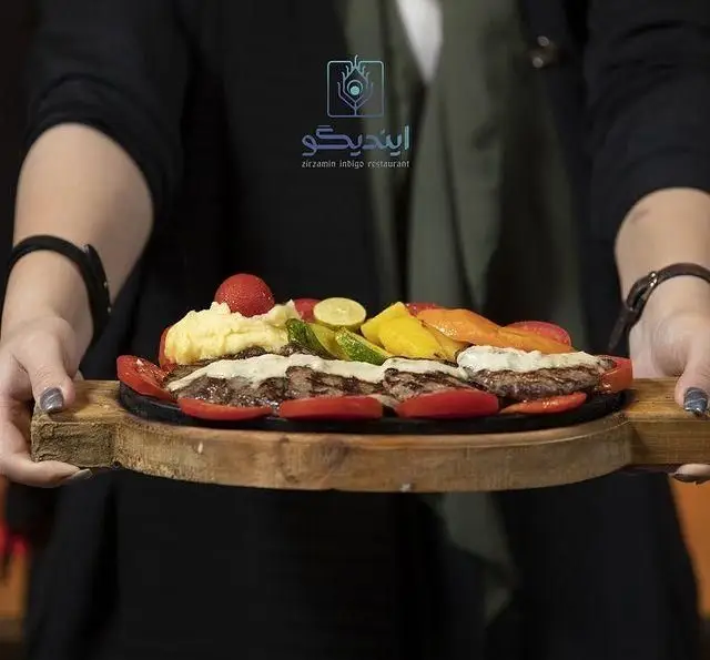 سرو غذای گرم و تازه کافه رستوران زیرزمین ایندیگو: بشقاب داغ استیک یا کباب با پوره سیب زمینی، گوجه و سبزیجات گریل شده روی سینی چوبی.