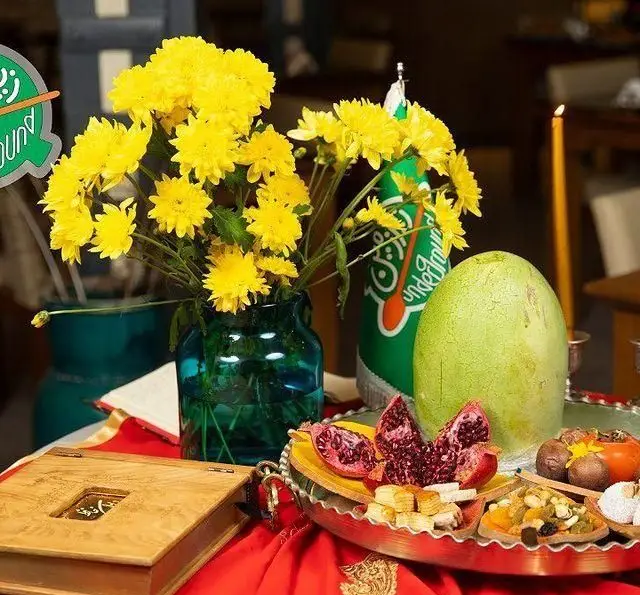 سفره شب یلدای کافه رستوران زیرزمین با چیدمان زیبا شامل گل‌های زرد، انار، هندوانه، آجیل و شیرینی برای دورهمی خانوادگی.