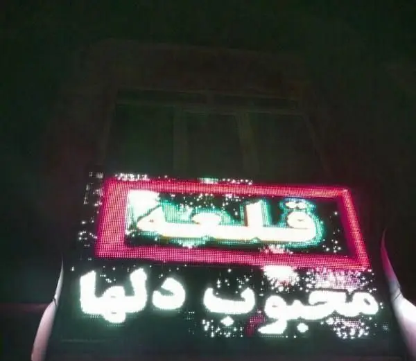 تصویری از نمای بیرونی فست فود ساندویچ قلعه در شب، با تابلوهای نئون درخشان. تابلوی بالایی نام "قلعة" را با نور سبز و پایینی "محبوب دلها" را نمایش می‌دهد.