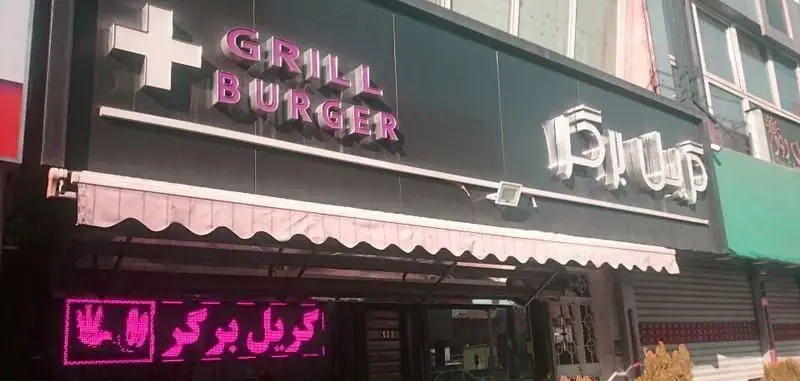 تابلوی مغازه فست فود گریل برگر با حروف برجسته صورتی و نماد به علاوه سفید، مکانی عالی برای تجربه طعم لذیذ همبرگر و ساندویچ.