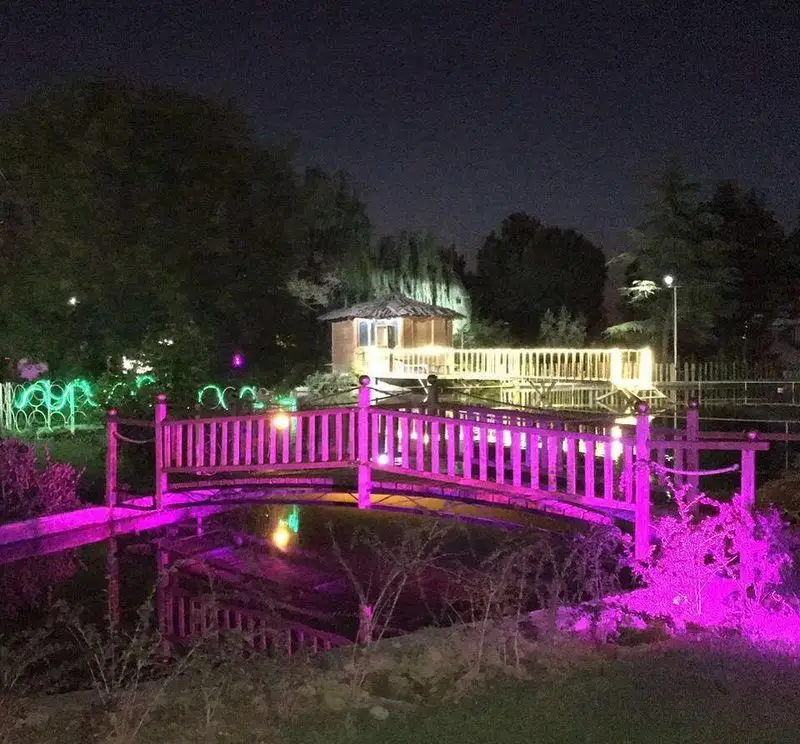 فضای شبانه و زیبای رستوران باغ گلستان با پل چوبی نورانی صورتی رنگ بر روی آب و آلاچیق روشن، محیطی دلنشین و رمانتیک.