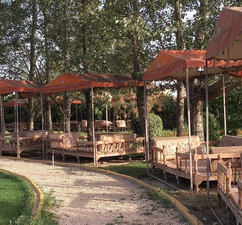 فضای دلپذیر و سنتی رستوران باغ گلستان با تخت‌های چوبی و کوسن، آلاچیق‌های نارنجی و مسیر شن‌ریزی شده میان درختان.