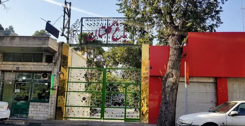 ورودی رستوران باغ گلستان با درب فلزی سبز رنگ و تزئینات زیبا، تابلوی نام در بالا و فضای سبز داخلی نمایان است.