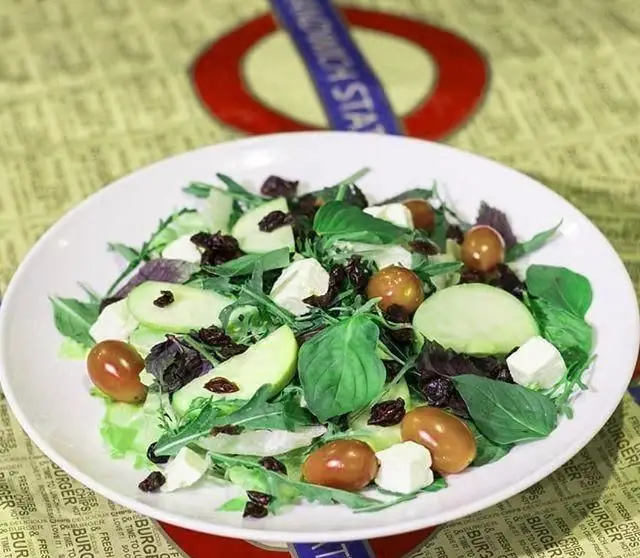 سالاد تازه و جذاب ساندویچی ایستگاه ساندویچ شامل برگ‌های سبز، سیب سبز، پنیر فتا، گوجه‌های گیلاسی و کشمش، گزینه‌ای سالم و خوشمزه.