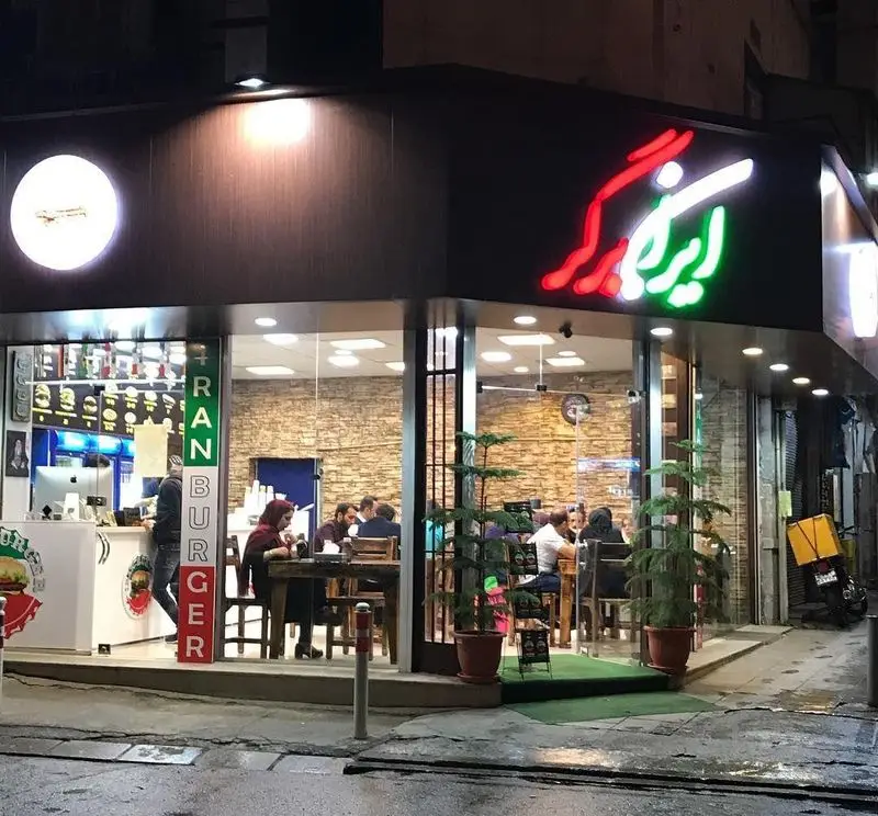 نمای بیرونی رستوران فست فود ایران برگر در شب با تابلوهای نئونی روشن و مشتریان در حال غذا خوردن داخل؛ مکانی جذاب.