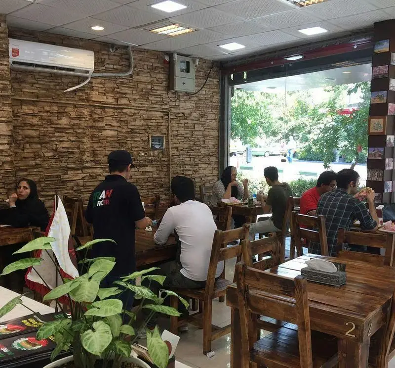 ایران برگر: فضایی دوستانه و دلپذیر با میز و صندلی‌های چوبی، مشتریان در حال صرف برگر و ساندویچ. پرسنل در حال سرویس‌دهی سریع و با کیفیت.