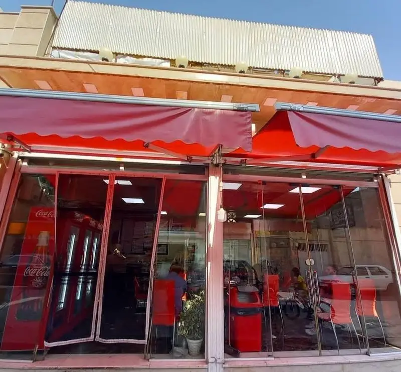تصویری از نمای خارجی و داخلی فست فود کوروش با سایبان‌های قرمز، میز و صندلی‌های شیک و لوگوی کوکاکولا، محیطی جذاب برای مشتریان.