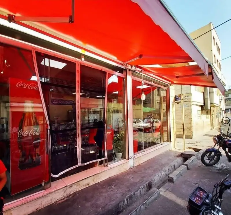 نمای بیرونی فست فود کوروش، رستورانی با سایبان‌های قرمز، درب‌های شیشه‌ای، میز و صندلی‌های قرمز و یخچال بزرگ کوکاکولا در یک خیابان شلوغ.