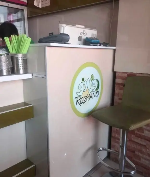 نمایی از پیشخوان فست فود روژانو با لوگوی بزرگ برند، دستگاه کارتخوان، سیستم POS و نی‌های سبز آماده پذیرایی از مشتریان