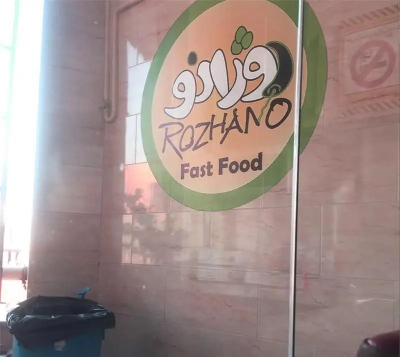 لوگوی فست فود روژانو با نام "ROZHANO Fast Food" روی تابلوی دیواری، شامل نوشته فارسی و انگلیسی با پس زمینه دیوارهای کاشی‌کاری شده.