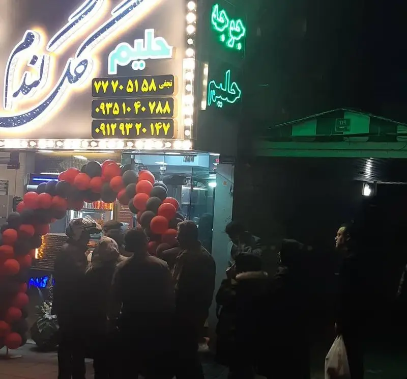تصویر شب از کبابسرای سنتی گل گندم با تابلوی نئونی حلیم و جوجه، جمعیت مشتریان در حال انتظار و تزئینات بادکنکی.