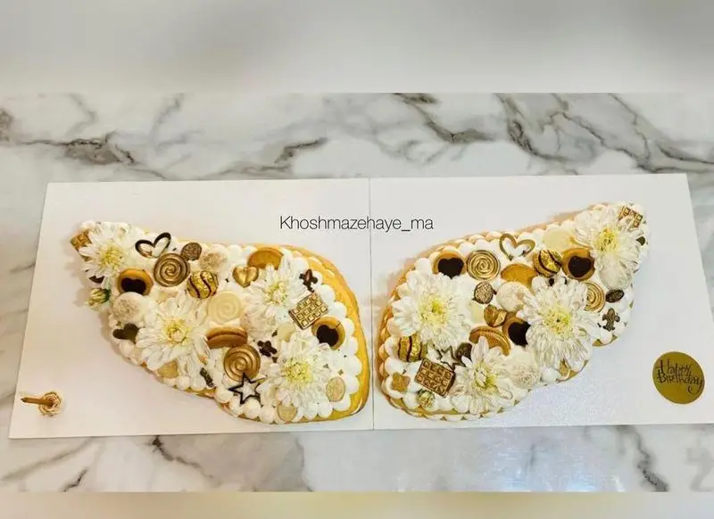 کیک کرم تارت زیبا و خوشمزه به شکل بال، تزئین شده با گل‌های سفید و شکلات‌های طلایی، مناسب جشن تولد و سفارش از "خوشمزه های ما".