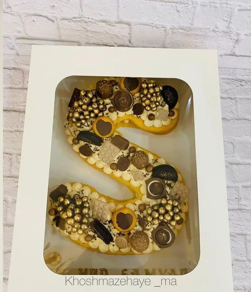 کیک حرف S سفارشی "خوشمزه های ما" با تزئین شکلات، مروارید طلایی و شیرینی‌های متنوع در جعبه شیک برای جشن و هدیه.