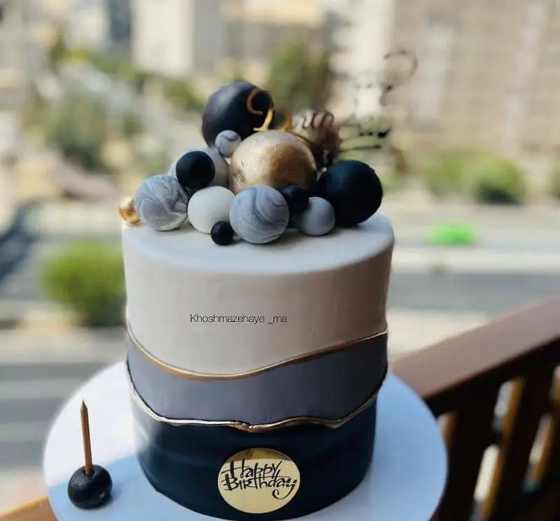 کیک تولد شیک و مدرن با تم رنگی مشکی، طوسی و سفید، تزئین شده با گوی‌های طلایی و مرمری و پلاک Happy Birthday. سفارش از خوشمزه‌های ما.