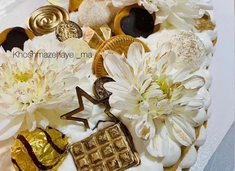 کیک خاص و لوکس تزیین شده با گل‌های طبیعی سفید و جزئیات طلایی، شکلات‌ها و بیسکوییت‌های متنوع، آماده سفارش از "کیک وشیرینی خوشمزه های ما".