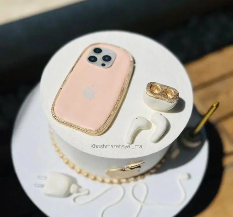 کیک تولد سفید دو طبقه با تزیینات گوشی آیفون صورتی، ایرپاد، شارژر و ایرفون. کیک سفارشی خوشمزه های ما.