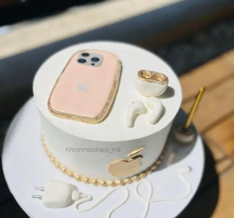 کیک تولد لاکچری با دیزاین گوشی آیفون، ایرپاد و لوگوی اپل در رنگ‌های سفید و طلایی، اثری خاص و شیک از "خوشمزه‌های ما".