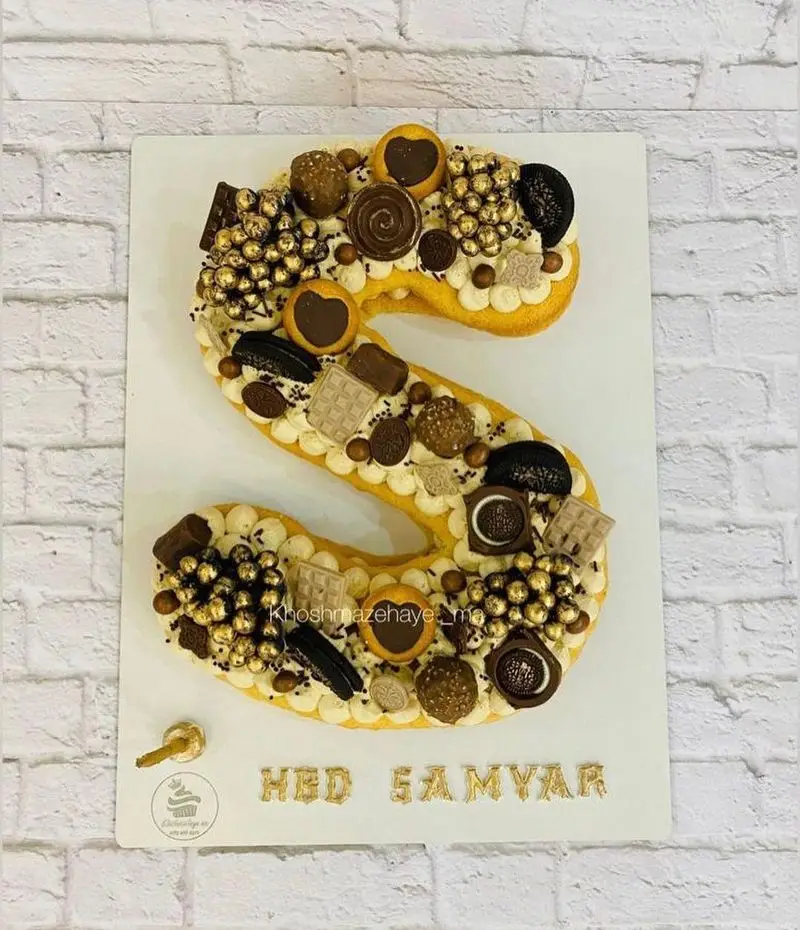 کیک حرف S شکلاتی و طلایی لوکس، تزئین شده با انواع کوکی و خوراکی‌های خوشمزه، از کیک و شیرینی خوشمزه های ما.