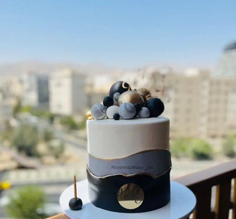 کیک مدرن و شیک خوشمزه های ما با دیزاین سه طبقه مشکی طوسی سفید و گوی‌های طلایی، مرمری، مناسب سفارش آنلاین انواع جشن و مناسبت.