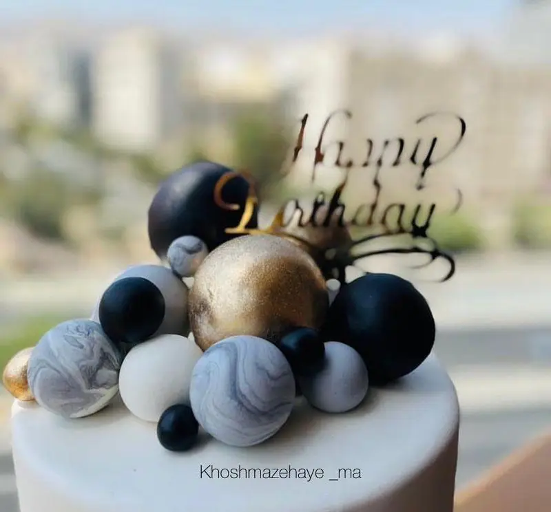 کیک تولد جذاب و سفارشی "خوشمزه های ما" با تزئین هنرمندانه گوی‌های مات و براق مشکی، طلایی، سفید و مرمری، برای لحظات خاص شما.