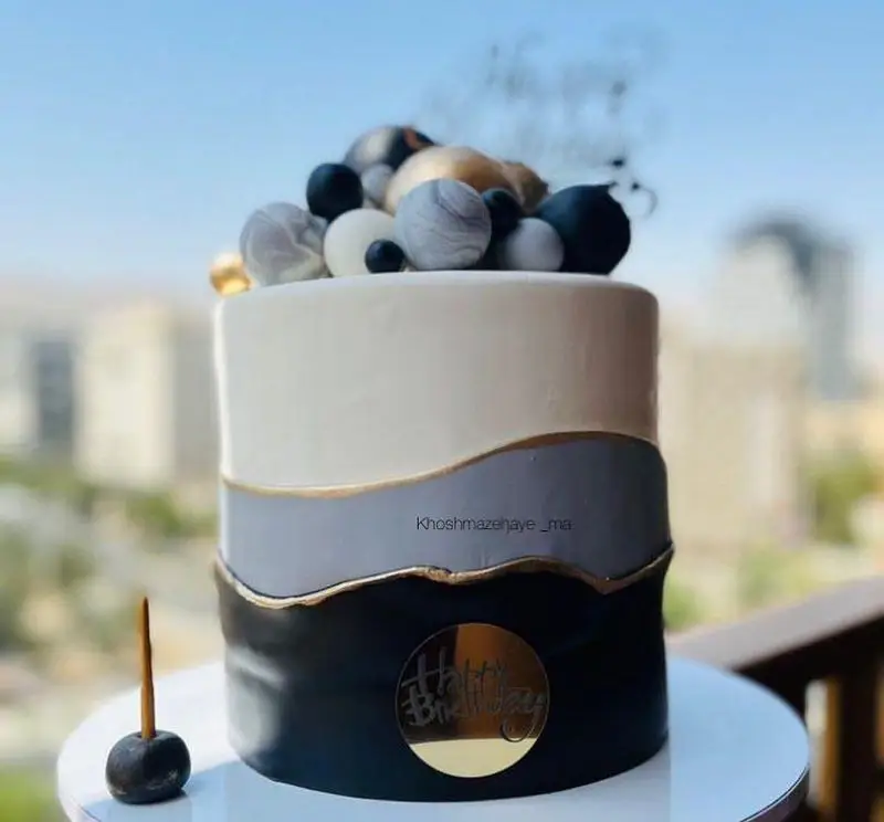 کیک تولد مدرن و شیک با دیزاین سه رنگ مشکی، طوسی و سفید، با نوارهای طلایی و گوی‌های دکوراتیو، مناسب جشن‌های خاص "خوشمزه‌های ما".