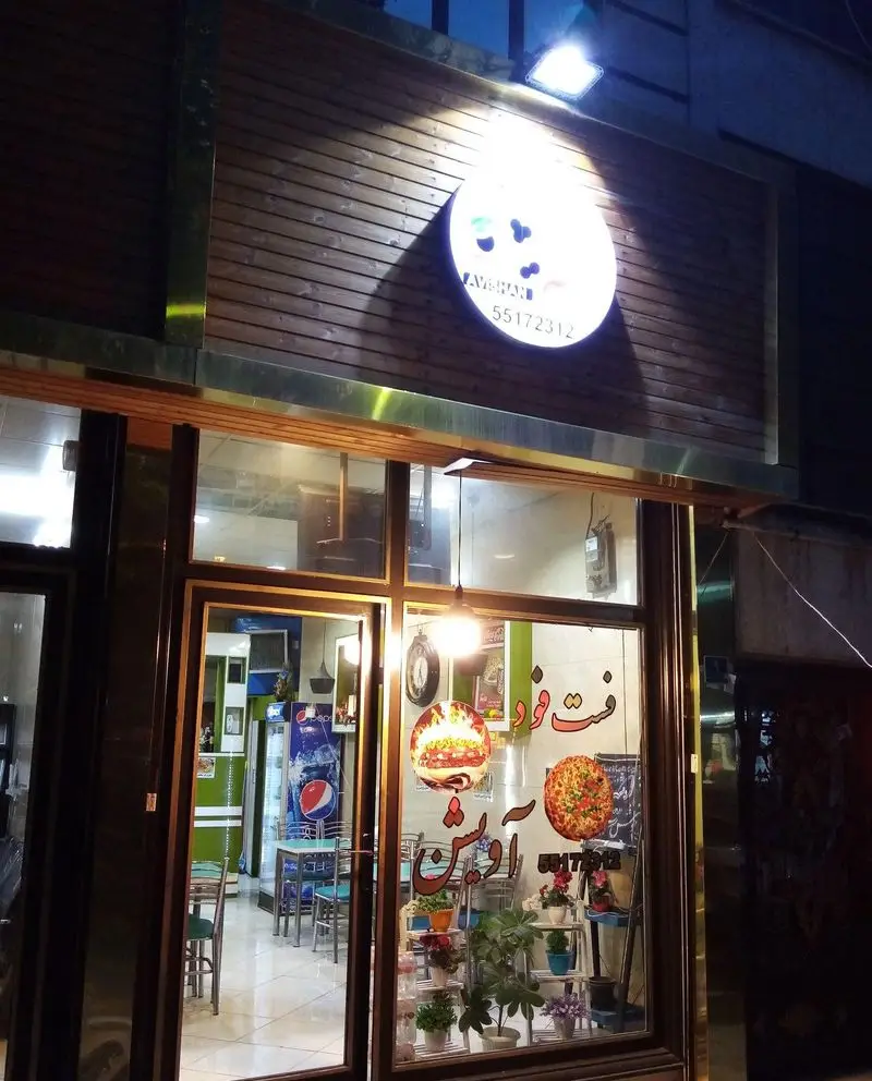 نمای شبانه فست فود پیتزا آویشن با تابلوی روشن، فضای داخلی دلنشین، میز و صندلی، یخچال نوشیدنی و گل و گیاه. منوی متنوع پیتزا و برگر.