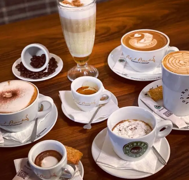 مجموعه‌ای از قهوه‌های لذیذ کافه روبرت: لاته، اسپرسو، کاپوچینو با طرح‌های هنری، کنار دانه قهوه روی میز چوبی.