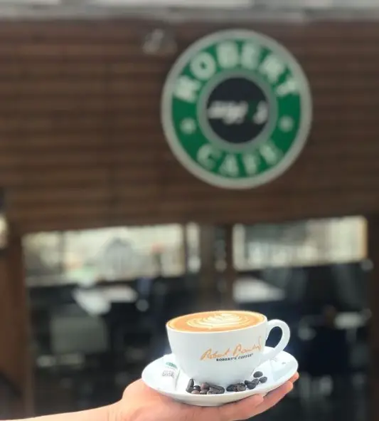 فنجان قهوه لاته با طرح هنری دلنشین در دست، در محیط دلپذیر کافه روبرت، با نشان تجاری کافه در پس زمینه.