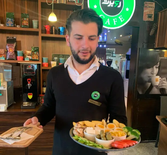 خدمتکار کافه روبرت در حال سرو یک سینی کامل و تازه صبحانه با تخم مرغ، پنیر و سبزیجات متنوع در فضای دلنشین کافه