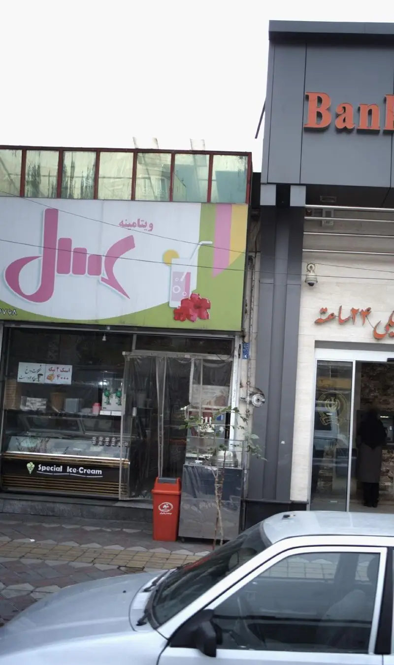 نمای بیرونی آبمیوه و بستنی فروشی ویتامینه عسل با تابلوی صورتی، پیشخوان بستنی و سطل زباله قرمز در پیاده‌رو شلوغ.