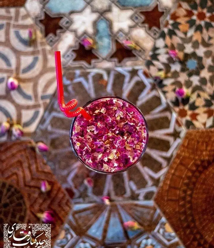 نوشیدنی سنتی گل محمدی با نی قرمز روی میز کاشی‌کاری شده در رستوران رفتاری. طرح‌های ایرانی زیبا و گلبرگ‌های پراکنده.