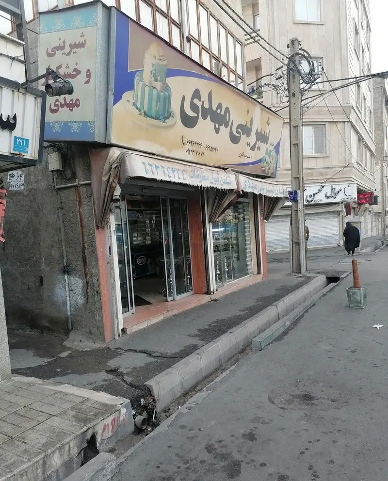 تصویر نمای بیرونی شیرینی سرای مهدی، قنادی و شیرینی‌فروشی با انواع کیک، شیرینی تر و خشک در یک خیابان شلوغ شهری.