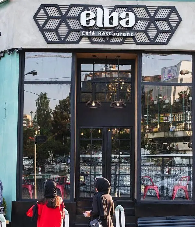 کافه رستوران البا: نمای خارجی شیک و مدرن، با پنجره‌های بزرگ و ورودی شیشه‌ای، نمایشگر فضای داخلی و خیابان شلوغ.