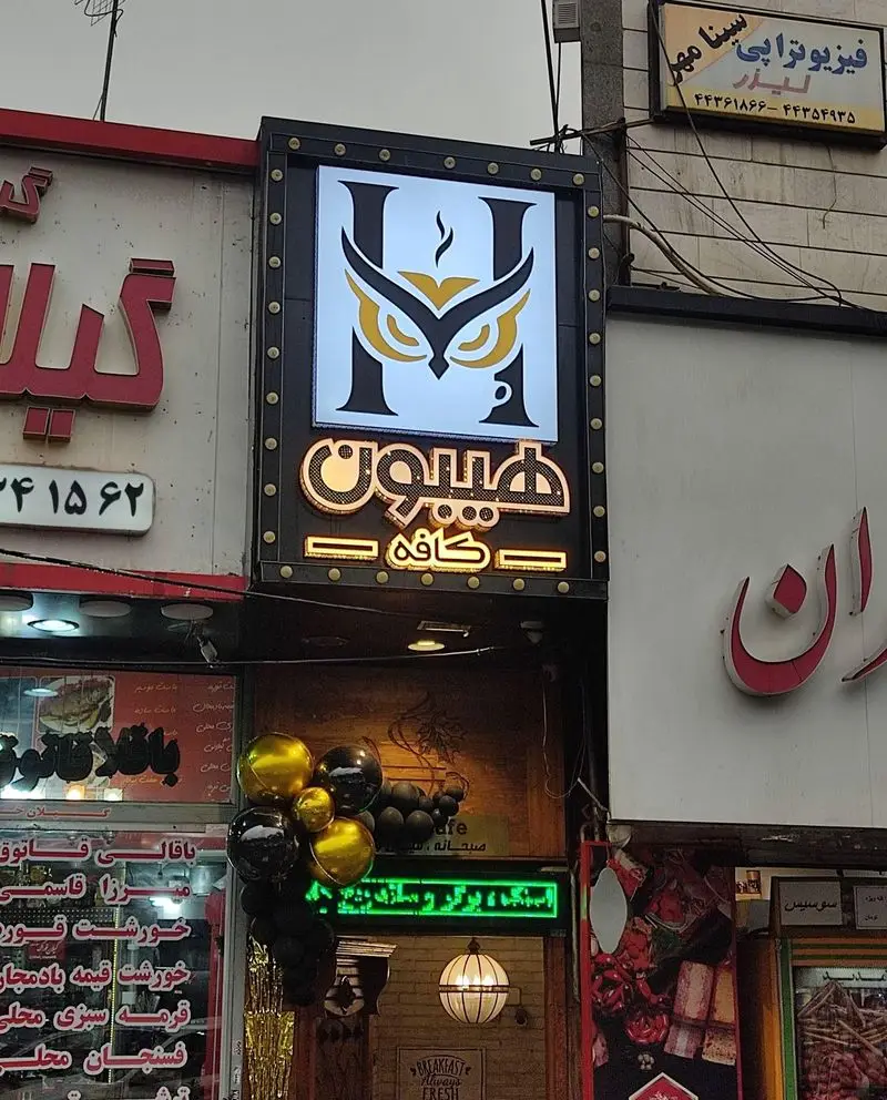 کافه هیبون: تابلوی نئونی درخشان با لوگوی خلاقانه جغد که در حرف H ادغام شده، نمای بیرونی این کافه را در خیابان به تصویر می‌کشد.