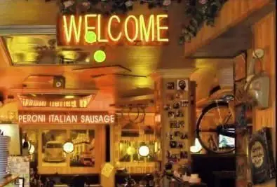 فضای داخلی دلنشین رستوران ایتالیایی رز ایتالیایی با تابلوی نئون "WELCOME" و "PERONI ITALIAN SAUSAGE" و دکوراسیون گرم و سنتی.