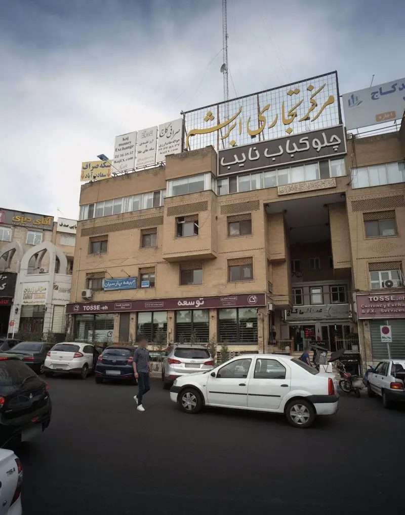 تصویر نمای بیرونی ساختمان مرکز تجاری ارشم، شامل رستوران چلو کباب نایب، صرافی کاج و بیمه پارسیان در خیابان‌های تهران.