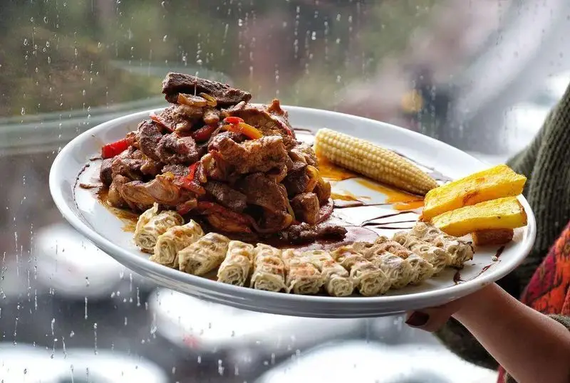 غذای لذیذ چلو کباب نایب: بشقاب چلوگوشت پر و پیمان با ذرت، مخلفات و رول‌های خاص در پس‌زمینه بارانی. طعم اصیل ایرانی.