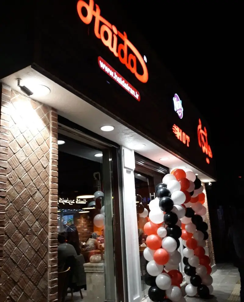 تصویر شب از نمای بیرونی شعبه ساندویچ هایدا هروی با تابلوی نورانی، بالون‌های افتتاحیه و آماده پذیرایی از مشتریان.