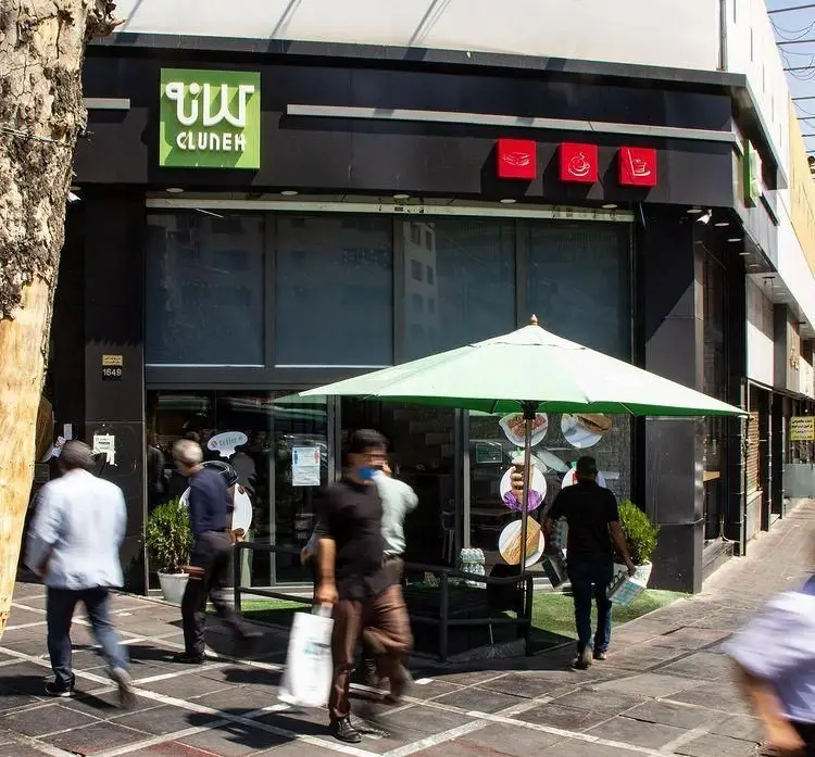 نمای بیرونی شعبه کافه رستوران کلانا با تابلوی سبز CLUNEH، چتر آفتابی و لوگوهای مربوط به غذا و نوشیدنی، همراه با عابرین.