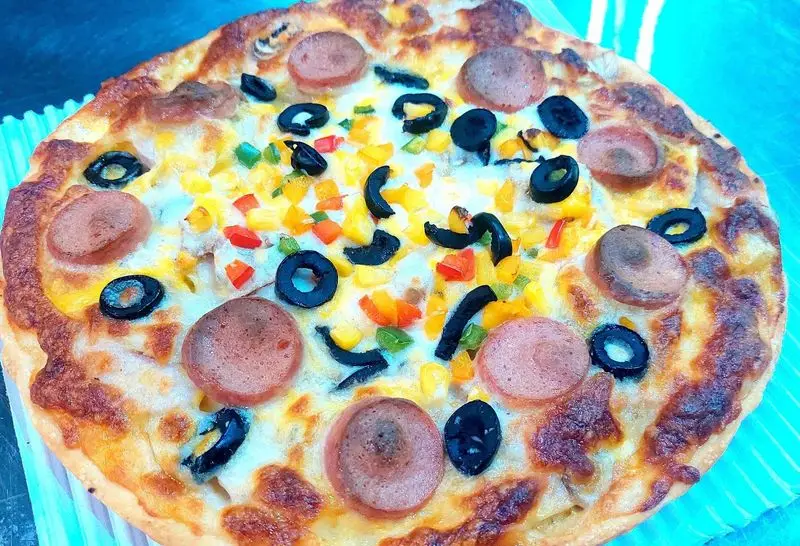 پیتزا سوسیس و زیتون با فلفل دلمه‌ای‌های رنگی (قرمز، زرد، سبز) و ذرت روی پنیر آب شده، محصول لذیذ فست فود آرام ناز.