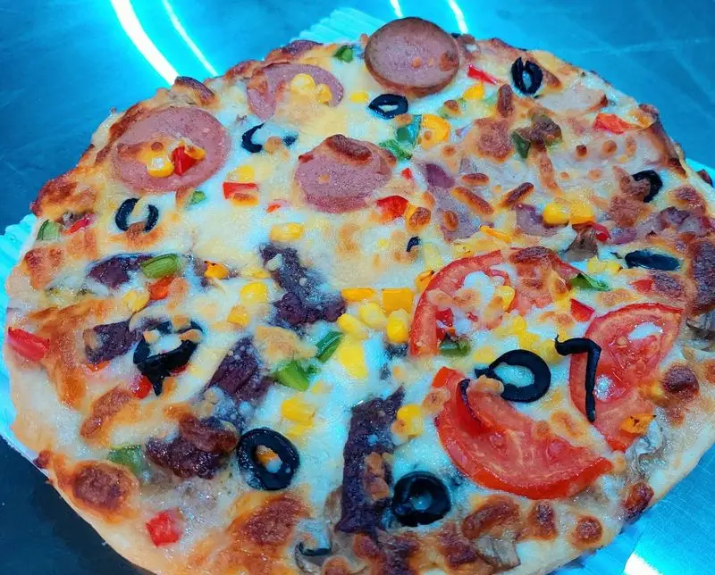 پیتزای خانگی و خوشرنگ از فست فود آرام ناز با ترکیب لذیذ پنیر، هات داگ، ذرت، زیتون سیاه، فلفل دلمه ای رنگی و گوجه فرنگی تازه.