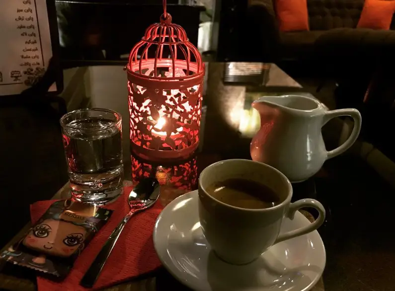 میز کافه کلبه در شب با اسپرسو، لیوان آب، فانوس شمعی زیبا و شکلات، برای لحظاتی دنج و آرام در فضایی گرم.