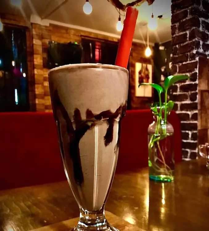 تصویری جذاب از میلک شیک شکلاتی کافه کلبه در محیطی دلنشین و دوستانه با دکوراسیون آجری و نورپردازی گرم، مکانی ایده‌آل برای لذت بردن.