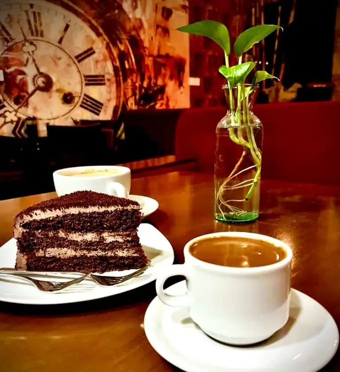 کافه کلبه تقدیم می‌کند: یک برش کیک شکلاتی لذیذ با لایه‌های کرمی و یک فنجان قهوه داغ، در فضایی دنج و دلپذیر با ساعت قدیمی و گلدان زیبا.