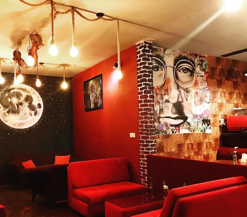 کافه کلبه: دکوراسیون داخلی هنری با نقاشی دیواری جان لنون و ماه، دیوارهای قرمز و آجری، مبلمان راحتی قرمز و نورپردازی گرم. فضایی دنج و مدرن.