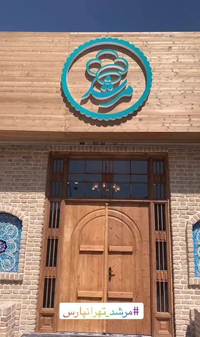 نمای شیک و سنتی رستوران مرشد در منطقه تهرانپارس با معماری آجری، درب چوبی نفیس و لوگوی آبی رنگ برند روی دیوار چوبی.