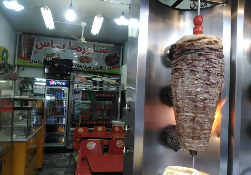 تصویر فست فود شاورما یاس با دستگاه بزرگ دونر گوشت و مرغ در حال آماده سازی، تابلوی منوی کباب ترکی و مرغ بریان، فضای داخلی با میز و صندلی.